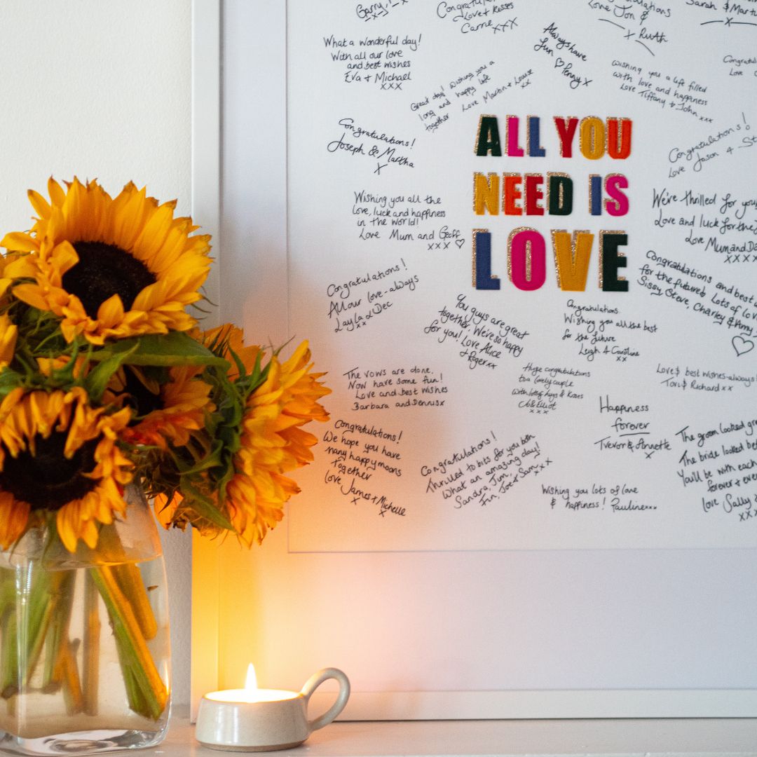 A handwritten Mr & Missive wedding guest book alternative featuring rainbow coloured letters and gold accents, shown with sunflowers and a lit candle in the foreground.