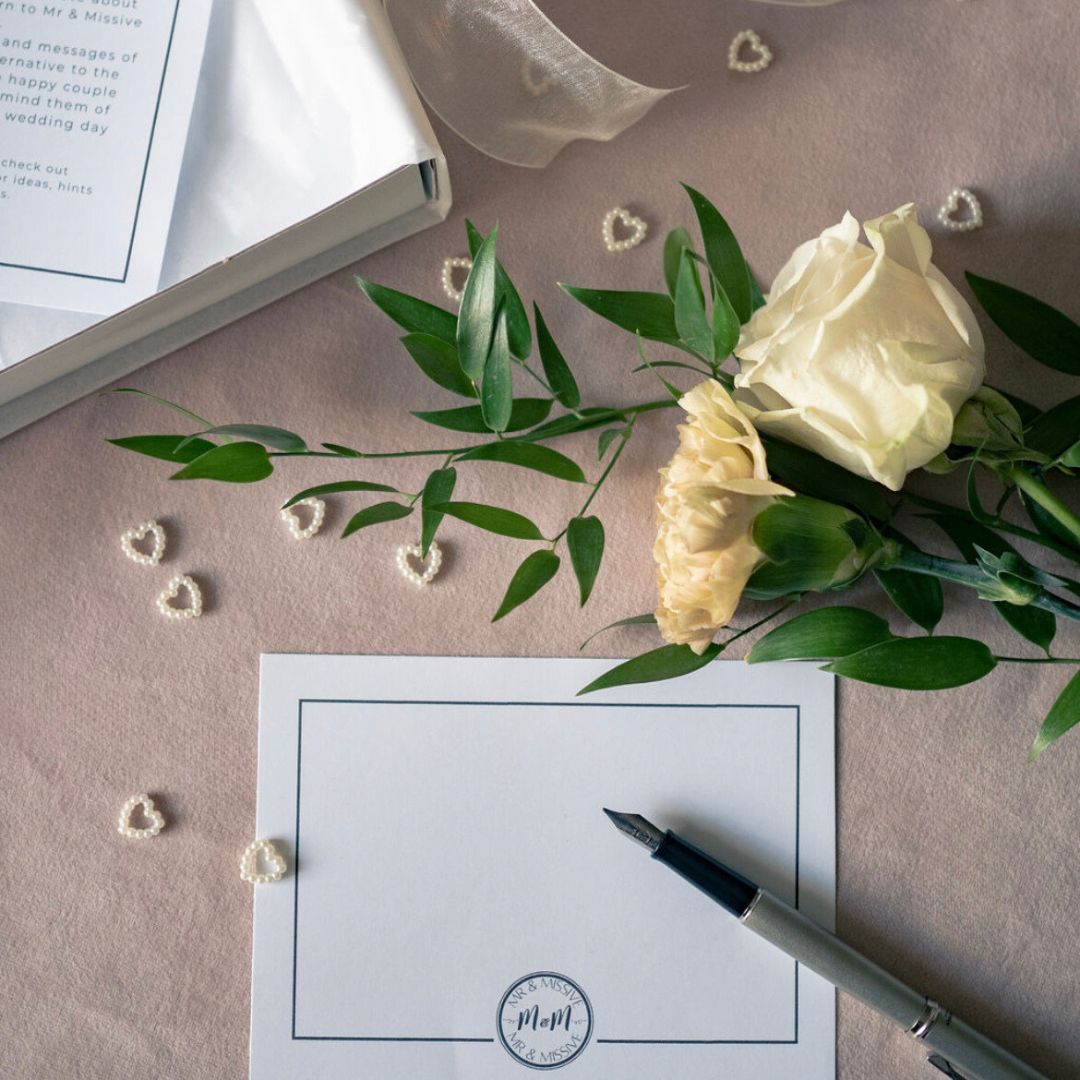 A flat lay of a Mr & Missive postcard for their wedding guest book alternative, shown with a fountain pen, wedding flowers and table décor.