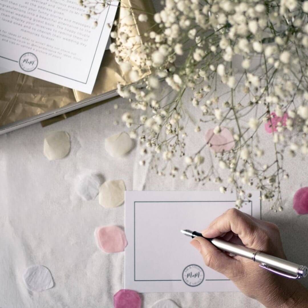 A flat lay of a wedding guest book alternative postcard with a hand holding the fountain pen with wedding table décor.